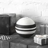 A black and white spherical vase is displayed on a textured surface in front of a white brick wall. Two gift-wrapped boxes are placed on either side of the vase, and a small black and white striped paper triangle is on the left. Náhľad obrázku produktu