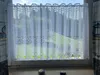 A white lace curtain is hung on a window, with a view of greenery and sky behind it. Two decorative stones are on the windowsill. Produktbild-Vorschau