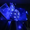 A white pineapple decoration is illuminated by blue LED string lights, placed on a dark fabric surface next to an open book. Produktbild-Vorschau