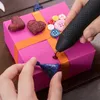 A person is using a black and pink glue gun to decorate a pink gift box with various buttons and heart-shaped embellishments. Produktbild-Vorschau
