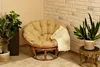 A papasan chair with a cushion and a throw blanket, placed next to two potted plants in a room with a brick wall. Produktbild-Vorschau