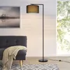 A floor lamp is placed next to a sofa in a living room setting, with a large window in the background. Produktbild-Vorschau