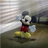 A Mickey Mouse plush toy is standing on a wooden floor, with light rays shining from the side. Voir les images du produit