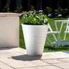 A white planter with flowers sits on a patio next to a wall, with a white table and chairs in the background. Voir les images du produit