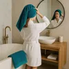 A woman in a white bathrobe is in a bathroom, wrapping a teal towel around her head. A teal towel is draped over the edge of a bathtub. Produktbild-Vorschau