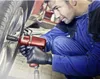 A mechanic in a blue uniform and black gloves is using a red impact wrench to tighten a lug nut on a car wheel. Produktbild-Vorschau