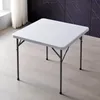 A white square folding table is shown in a room with light wood flooring and a dark patterned wall in the background. Produktbild-Vorschau