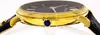 Close-up side view of a gold watch with a black face and a blue jewel on the crown. Produktbild-Vorschau