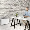 A man works on a laptop at a desk in front of a brick wall, with a white heater on the floor to the left. Anteprima dell’immagine prodotto