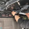 A person is working on the underside of a vehicle, possibly installing or inspecting something. Produktbild-Vorschau