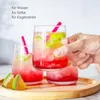 A hand holding a glass of pink beverage with lime slices and ice, with two other similar glasses in the background on a wooden tray. Produktbild-Vorschau