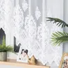 A white lace curtain with a damask pattern is shown hanging in front of a window. There are also some decorative items on the windowsill, including a framed photo and a small golden figurine. Náhled obrázku produktu