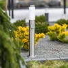 A silver bollard light with a white top is positioned in a garden setting, surrounded by gravel and greenery. Produktbild-Vorschau