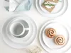 A neatly arranged set of white ceramic plates and a mug, with a folded napkin and a small sprig of flowers. Produktbild-Vorschau