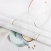 A close-up view of a beige roller blind with a decorative embroidered wave pattern, draped over a light blue cup of coffee and saucer on a white wooden surface. Produktbild-Vorschau