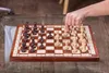 A hand is shown moving a chess piece on a wooden chessboard. Anteprima dell’immagine prodotto