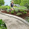 A woman is gardening in a backyard, tending to plants in a raised garden bed with a small greenhouse. Produktbild-Vorschau