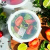A close-up overhead view of a round glass food container filled with salad, surrounded by fresh ingredients like tomatoes, lime, and parsley on a wooden cutting board. Produktbild-Vorschau