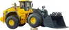 A yellow and gray toy construction vehicle, a Volvo wheel loader, is shown against a white background. The loader is positioned with its front bucket slightly raised and resting on a small rock. Náhľad obrázku produktu