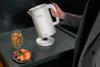 A person is holding a white electric kettle over a kitchen counter, with steam rising from the spout. A glass of tea and a croissant with berries are on the counter. Produktbild-Vorschau