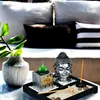 A zen garden set with a Buddha head, candle, and rake on a marble table, with pillows in the background. Náhled obrázku produktu