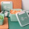Three green food storage containers, two filled with frozen food (peas and diced carrots), are arranged on a green surface with cork coasters and a striped towel. Náhľad obrázku produktu