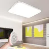 A hand holding a remote control in front of a modern living room with a ceiling light and artwork. Produktbild-Vorschau