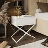 A white nightstand with a drawer is placed next to a bed in a bedroom. A lamp, books, and decorative items are on top of the nightstand. Dried plants are on the left, and a rug is on the floor. Produktbild-Vorschau