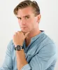 A man wearing a blue shirt and a watch, with his hand on his chin. Produktbild-Vorschau