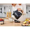 A person pouring hot water from a black electric kettle into a mug in a kitchen setting. Produktbild-Vorschau