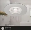 A close-up view of a white ceiling light fixture with a chrome trim, set against a blurred background of a bathroom. Produktbild-Vorschau