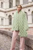 A woman stands on a stone walkway in front of a building, wearing a green textured cardigan and pink pants. Produktbild-Vorschau