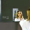 A woman is looking into a smart mirror that displays weather and time information. Produktbild-Vorschau