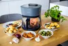 An air fryer is presented on a wooden table, surrounded by various prepared food items like roasted chicken, potatoes, sweet potato fries, Brussels sprouts, and mixed vegetables. A small potted plant is visible in the background. Produktbild-Vorschau