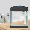 A small aquarium with a filter pump inside, placed on a wooden surface with a plant and a lamp in the background. Náhled obrázku produktu