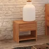 A wooden nightstand with a drawer and an open shelf, placed next to a bed. A white lamp sits on top of the nightstand. Produktbild-Vorschau