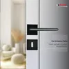 A close-up of a black door handle and lock mechanism on a white door, with a blurred background of a room interior. Produktbild-Vorschau
