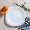 A white square plate with rounded corners is set on a textured light gray surface. Two halves of an orange are placed on the top left of the plate. A sprig of green leaves is to the top right of the plate, and a blue napkin with cutlery is to the bottom ri Náhled obrázku produktu