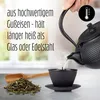 A hand pouring hot water from a cast iron teapot into a cast iron teacup, with loose tea leaves and a wooden scoop in the foreground. Produktbild-Vorschau