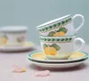 Two stacked teacups with saucers, decorated with a fruit and leaf pattern, are placed on a table. A few pink petals are scattered in the foreground. Produktbild-Vorschau