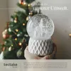 A hand holding a snow globe with a Christmas tree inside, with a blurred Christmas tree in the background. Produktbild-Vorschau