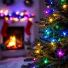 A close-up of a Christmas tree decorated with colorful lights, with a fireplace and stockings visible in the blurred background. Produktbild-Vorschau