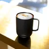 A black Ember mug filled with a frothy beverage sits on a golden surface, with soft light illuminating the scene. Náhled obrázku produktu