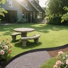 A garden scene with a stone picnic table and benches, a pathway with gravel, and roses in the foreground, with a house in the background. Náhled obrázku produktu