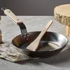 A frying pan with a wooden handle and a wooden spatula inside, placed on a textured surface. Anteprima dell’immagine prodotto
