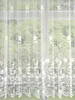 A white lace curtain with a floral pattern hangs in front of a window, obscuring the view of green trees outside. Produktbild-Vorschau
