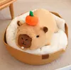 A plush capybara toy wearing a pumpkin hat, lying in a woven basket bed with a white cushion. Náhled obrázku produktu