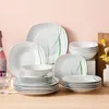 A collection of white porcelain dinnerware with green grass-like accents, arranged on a light brown surface against a light pink background. Náhled obrázku produktu