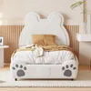 A white children's bed with a bear-ear shaped headboard and paw print details on the footboard, set in a bedroom with a beige fluted wall panel and a small white side table. Produktbild-Vorschau