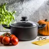 A black pot with a glass lid is placed on a white surface, surrounded by fresh ingredients like tomatoes, basil, spaghetti, and a pumpkin. The background is a textured gray wall with dramatic lighting. Náhled obrázku produktu